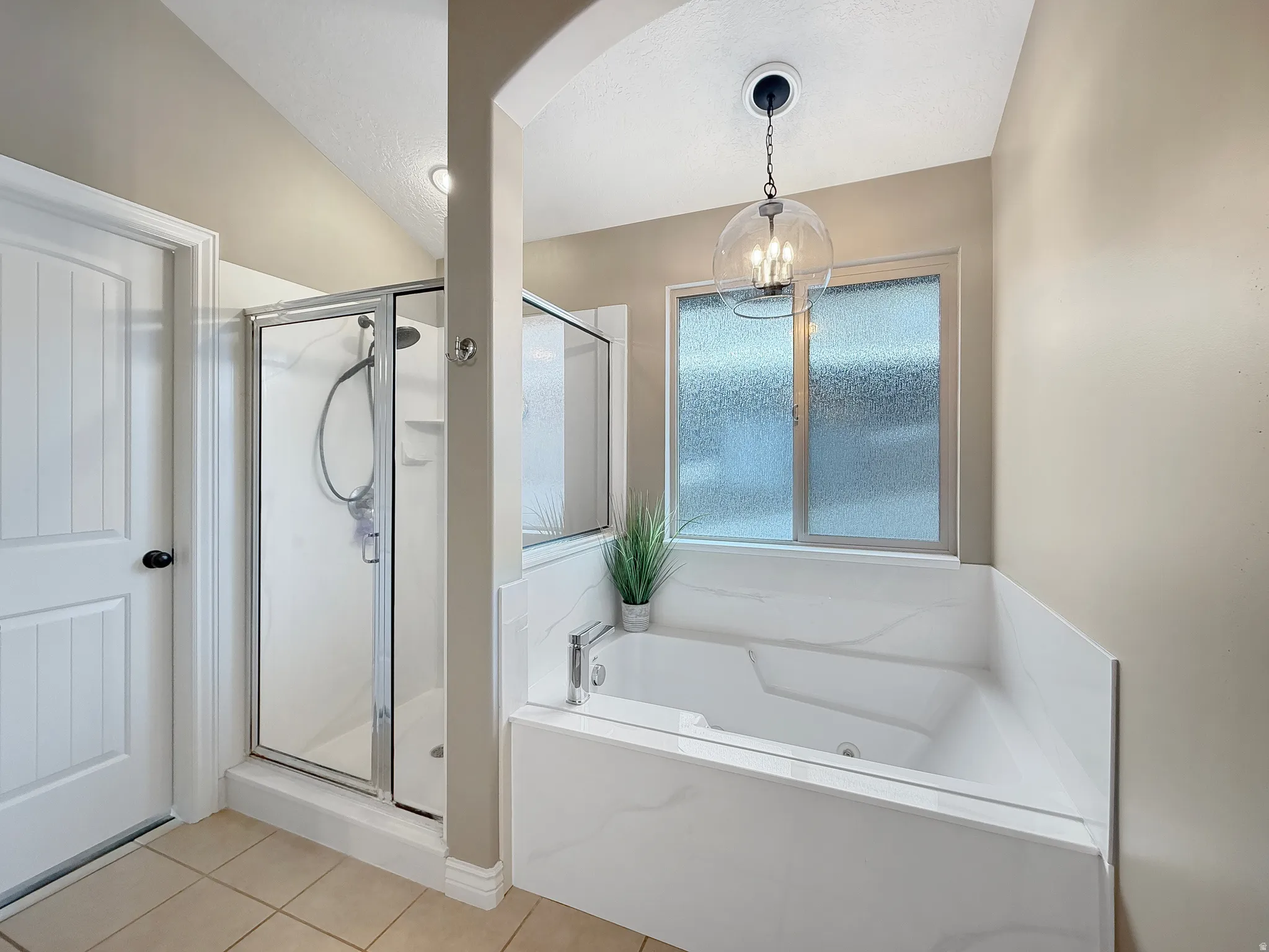 Tiled bathroom featuring a built-in soaking tub with white-patterned surround, a glass-enclosed shower, and a frosted window