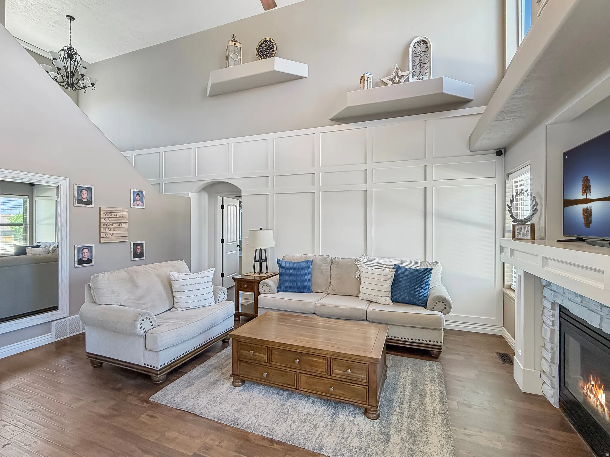 Living area with high ceilings, decorative wainscoting, wood-finish flooring, and a stone-surround fireplace