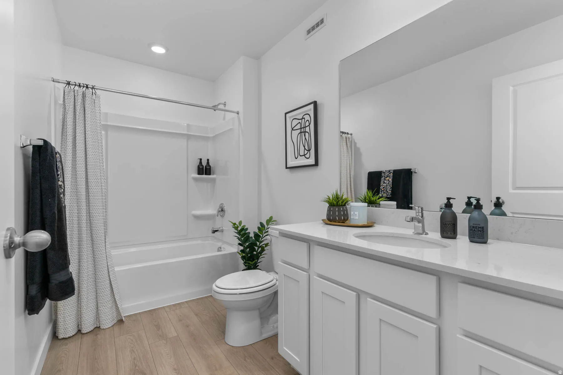 Bathroom featuring wood-finish flooring, a white vanity with a light-toned countertop, an integrated sink with a chrome faucet, and a combination tub and shower with white surround panels