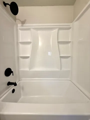 White bathtub and shower surround featuring integrated shelving, a matte black shower head, and matte black bath fixtures