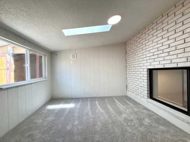 Spacious room featuring a white-painted brick accent wall with a rectangular fireplace, a large window, a skylight, and recessed lighting