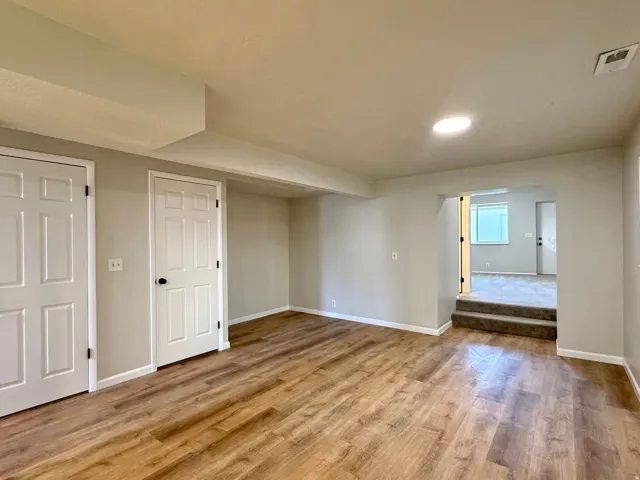 Spacious room featuring wood-finish flooring, two panel doors with black hardware, recessed ceiling lighting, and a stepped entry to an additional room with carpeting
