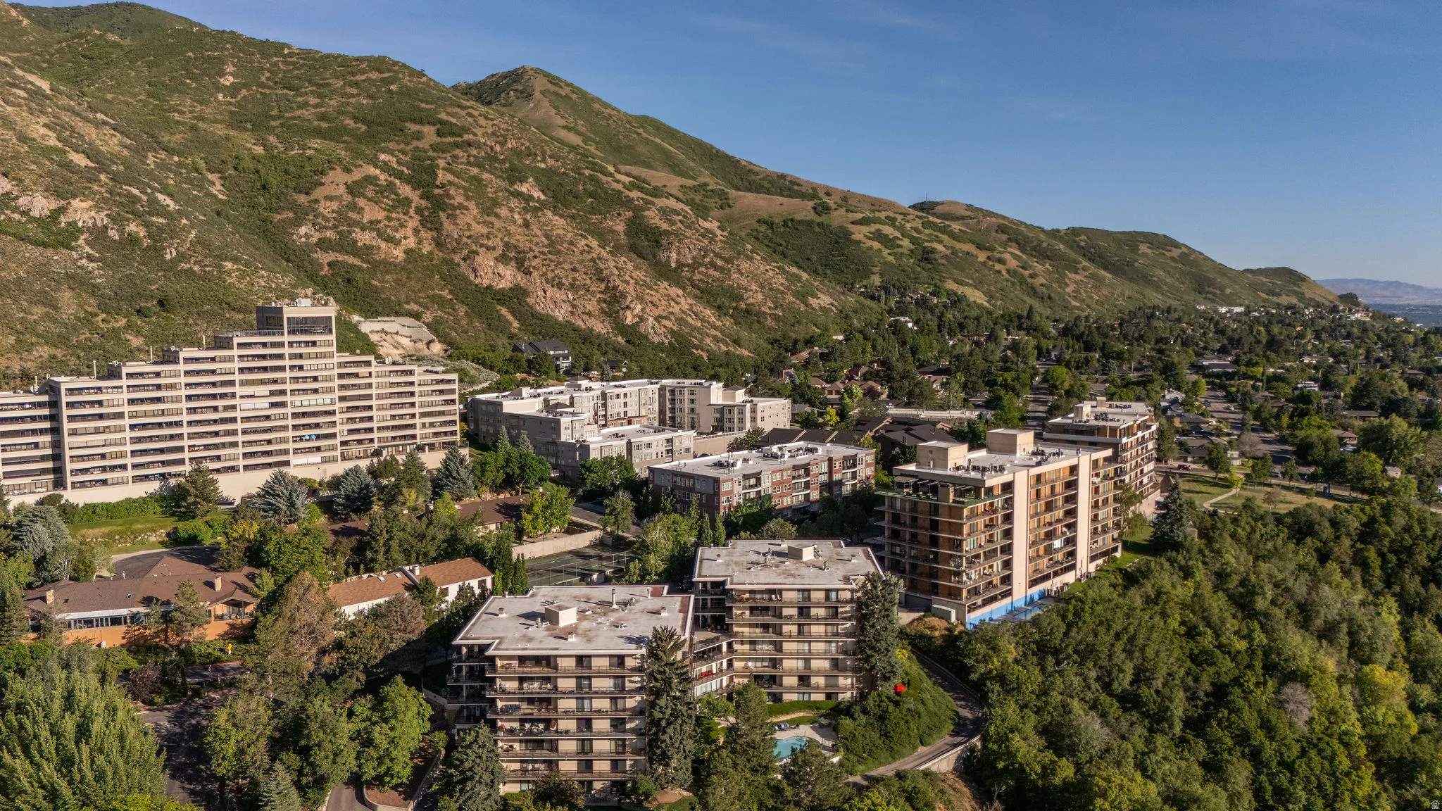 Terraced apartment buildings featuring multiple balconies, surrounded by lush green trees and natural mountain terrain