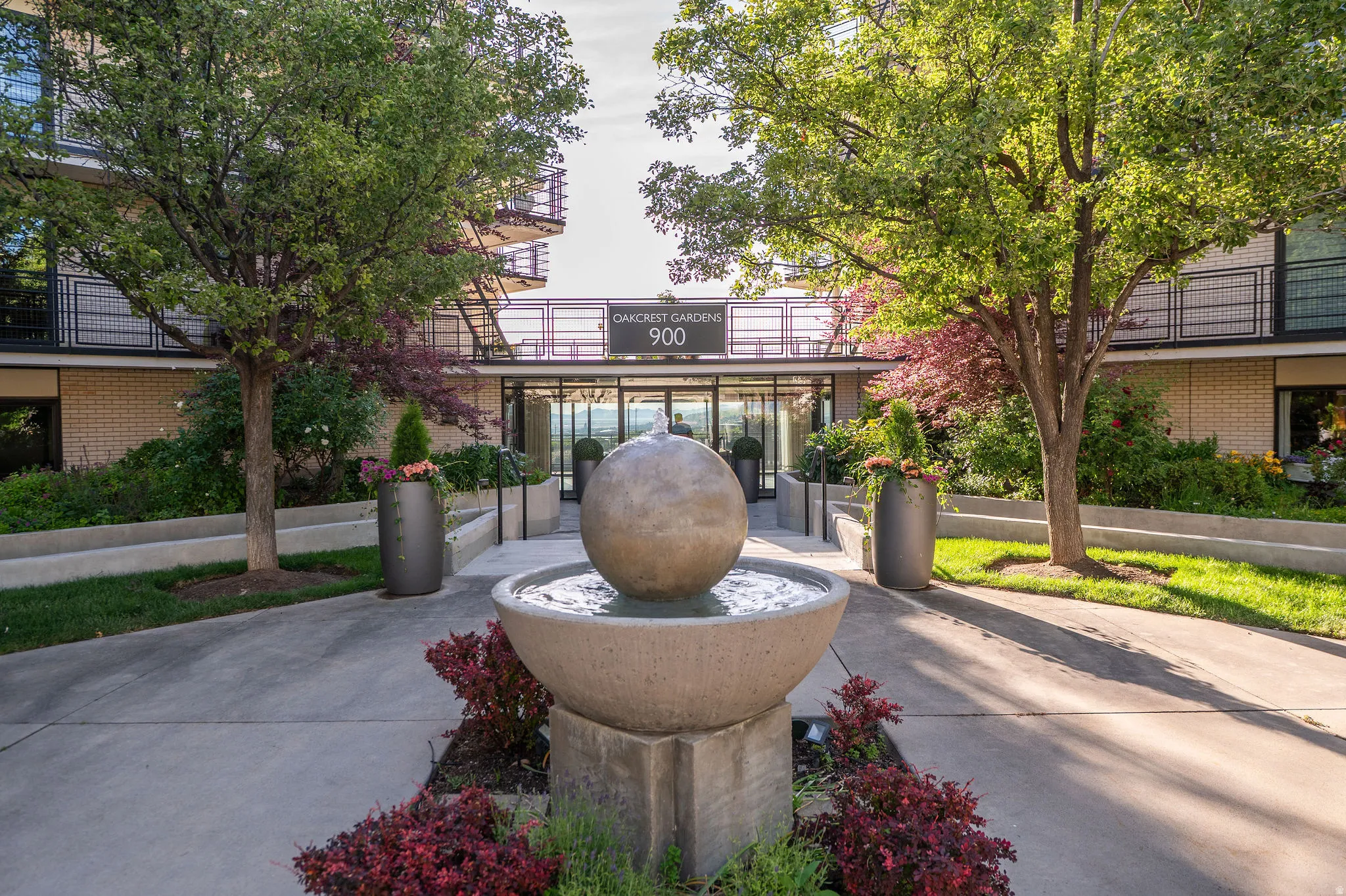 Contemporary building entrance featuring a spherical water fountain, lush plantings, mature trees, and a concrete pathway leading to glass entry doors