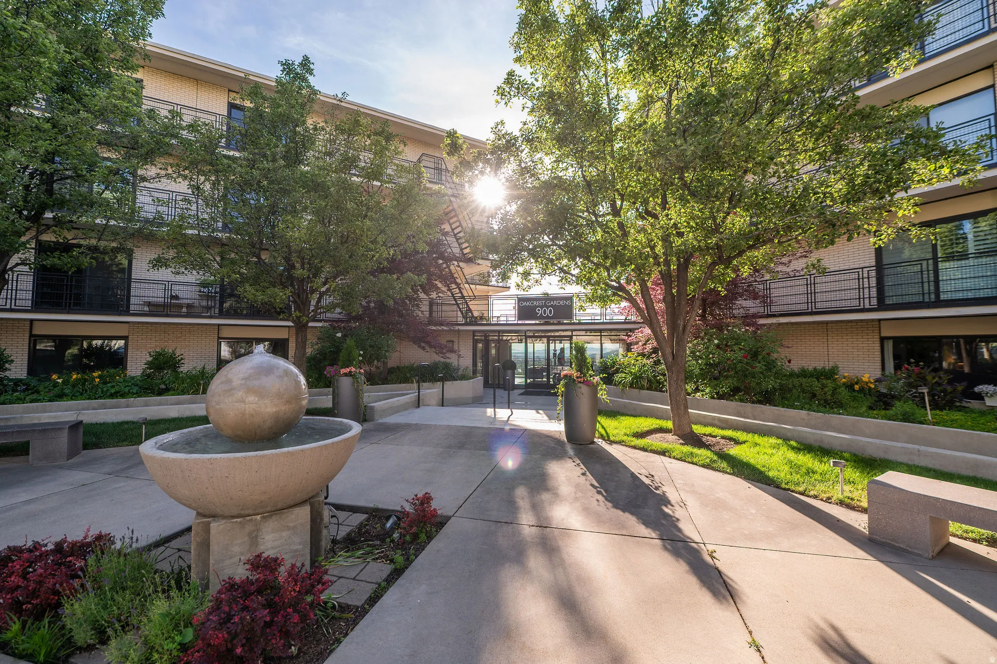 Building entrance featuring a contemporary fountain, manicured landscaping, concrete pathways, and built-in benches