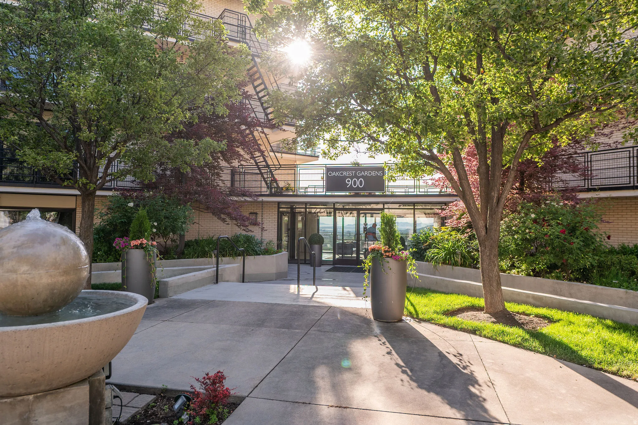 Building entrance featuring a contemporary spherical water feature, mature trees, and landscaping with varied plantings