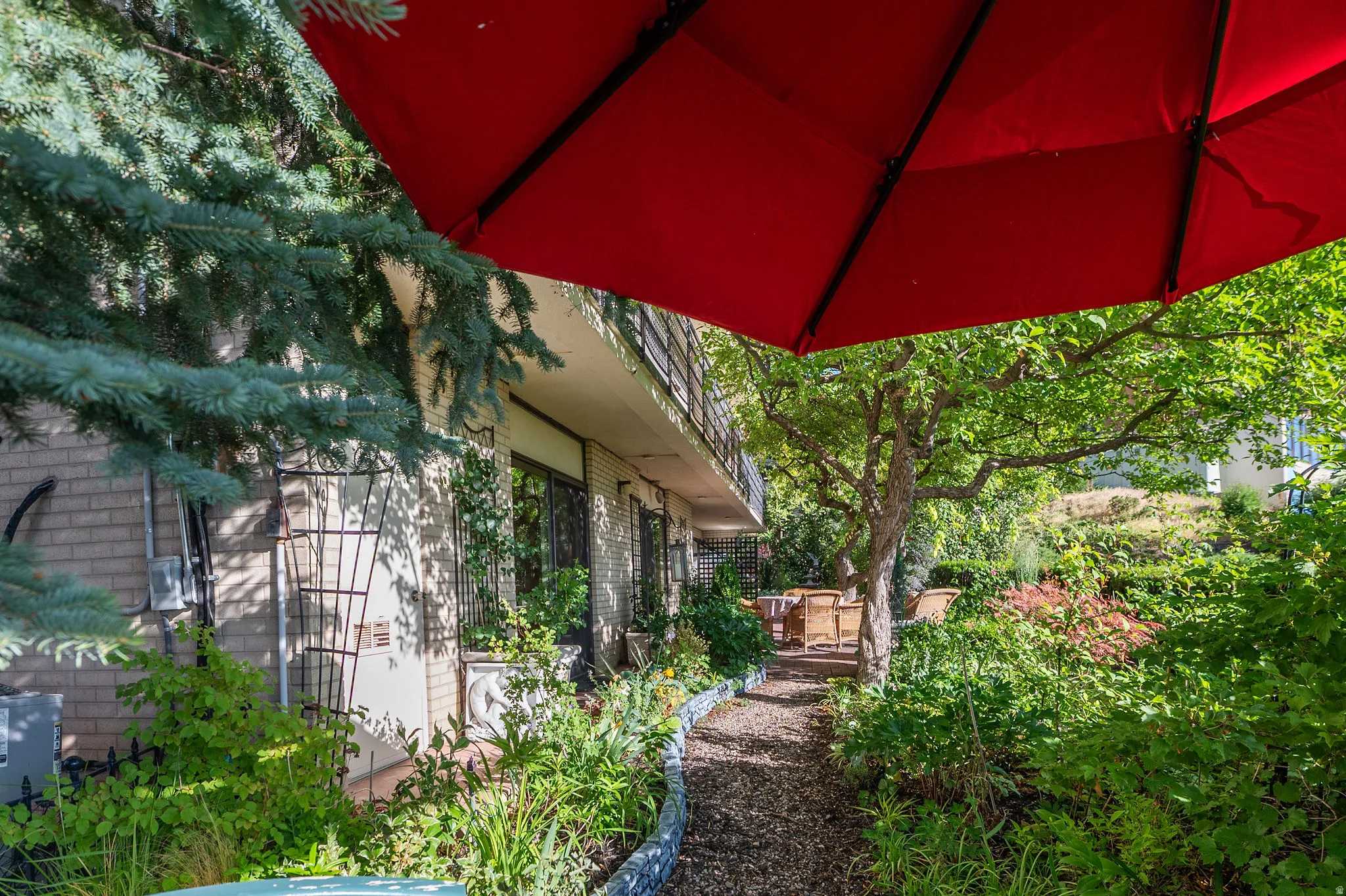 Lush outdoor area featuring a winding path, mature trees, dense plantings, and a brick-faced exterior with large windows