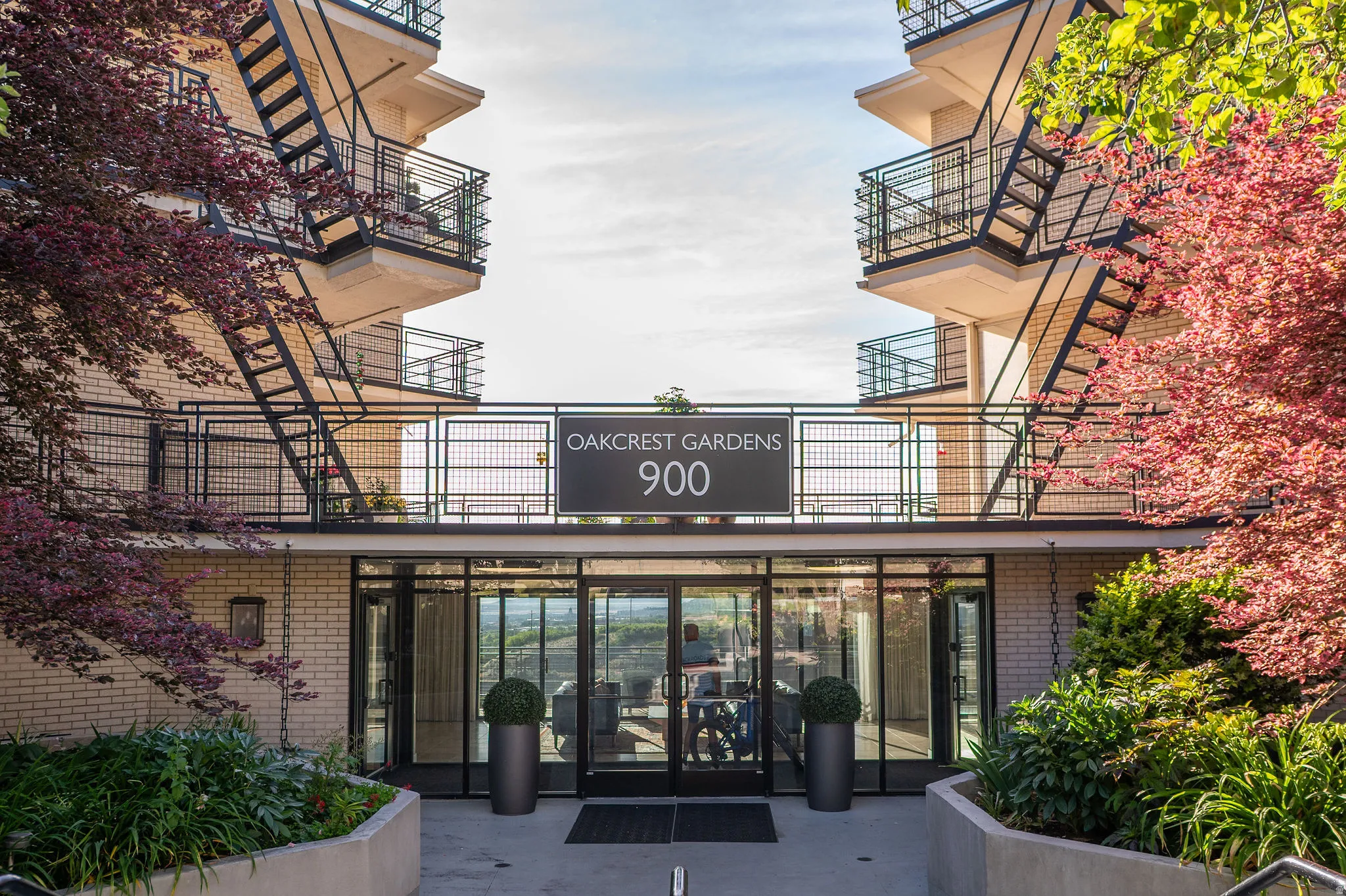 Building entrance featuring extensive glass doors, light-colored brick facade, black metal railings, and integrated landscaping with mature trees and shrubs
