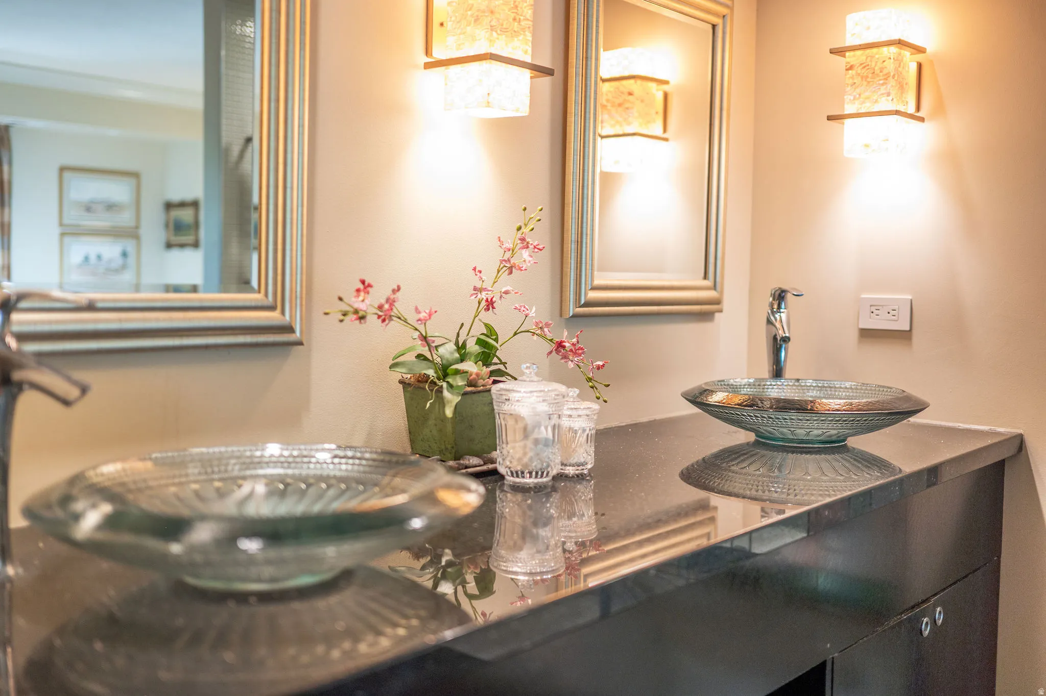 Dual glass vessel sinks set on a dark countertop with two framed wall mirrors and decorative wall sconces