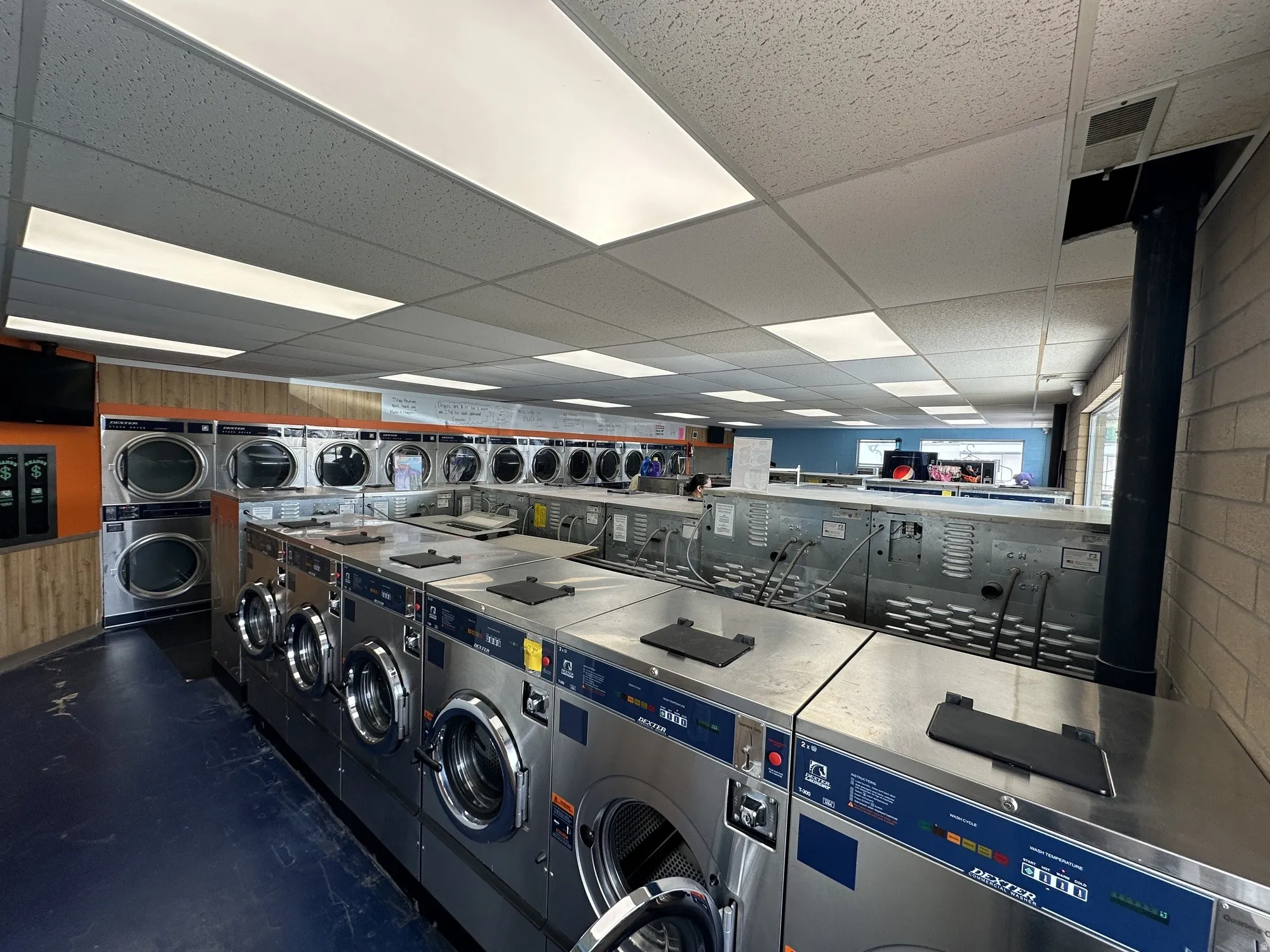 Commercial laundry facility featuring multiple front-load and top-load washing machines, a drop ceiling with integrated lighting panels, and durable block wall construction