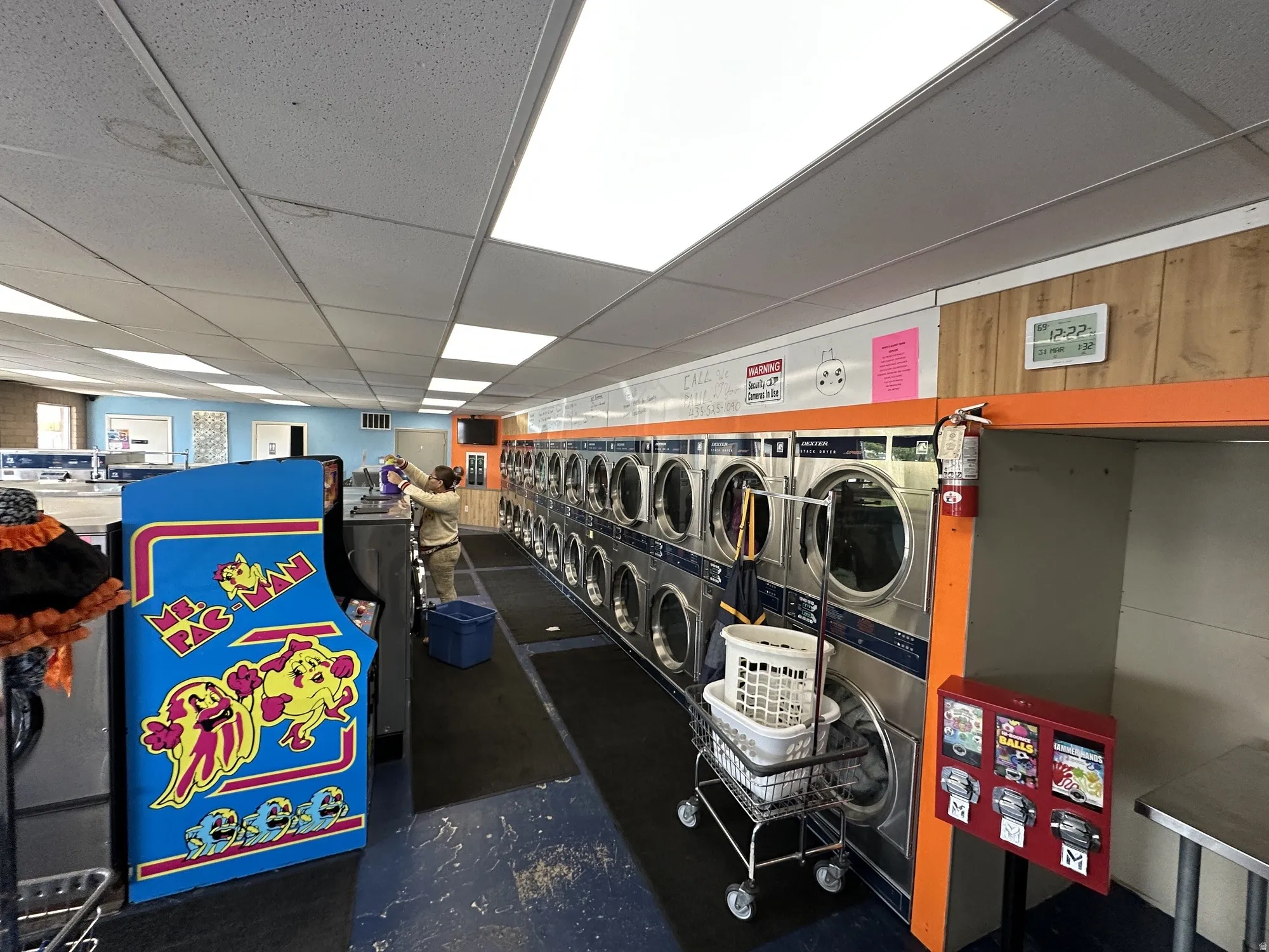 Commercial laundry facility featuring multiple stainless steel washing machines, drop ceiling with integrated fluorescent lighting, wood-paneled wall accents, and durable flooring