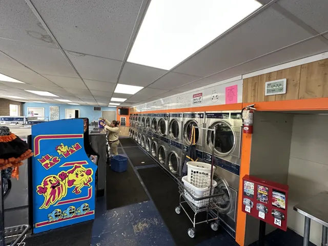 Commercial laundry facility featuring multiple stainless steel washing machines, drop ceiling with integrated fluorescent lighting, wood-paneled wall accents, and durable flooring