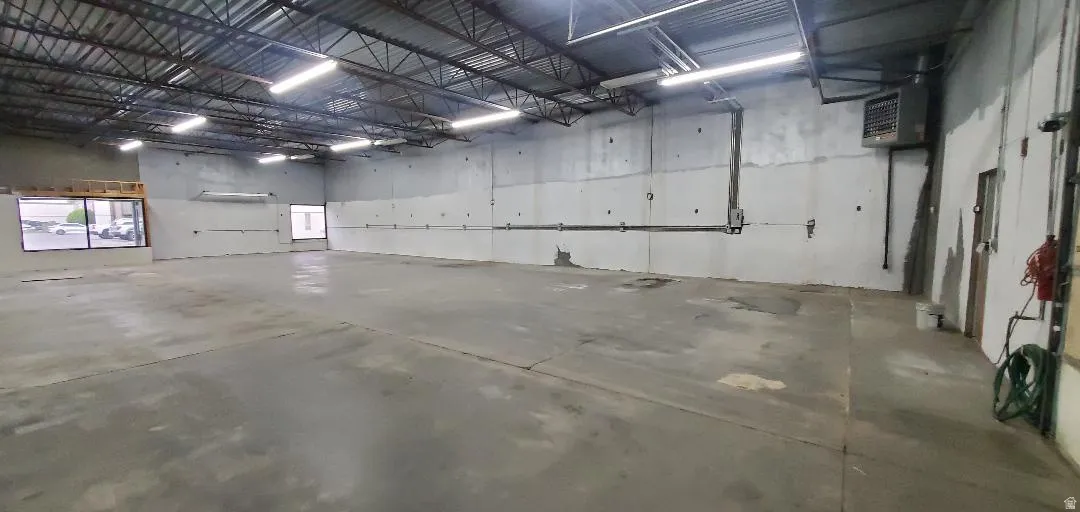 Expansive open warehouse space featuring concrete flooring, exposed ceiling trusses, abundant fluorescent lighting, multiple wall outlets, and large street-facing windows