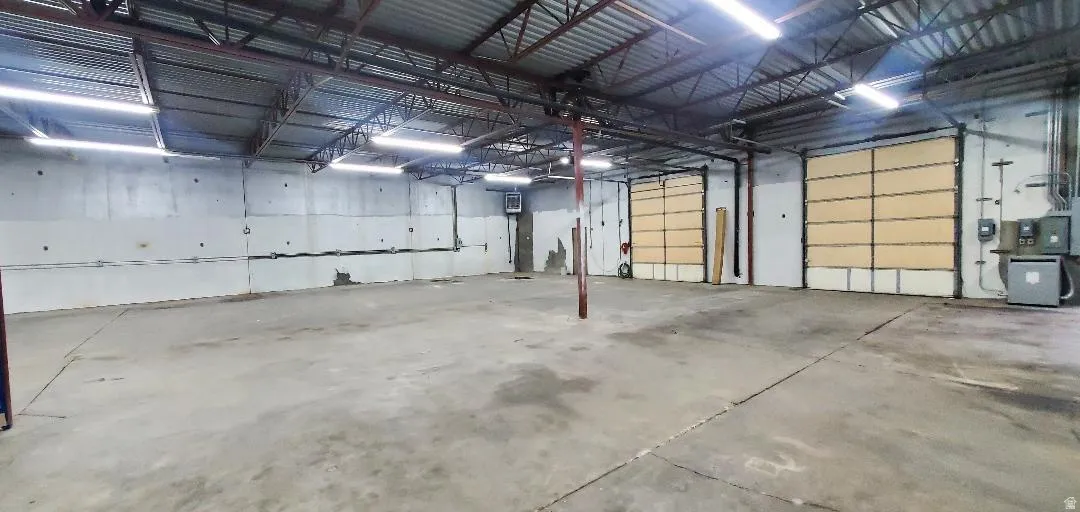 Spacious commercial interior featuring a concrete floor, exposed metal truss ceiling, fluorescent lighting, two large roll-up doors, and exposed block walls