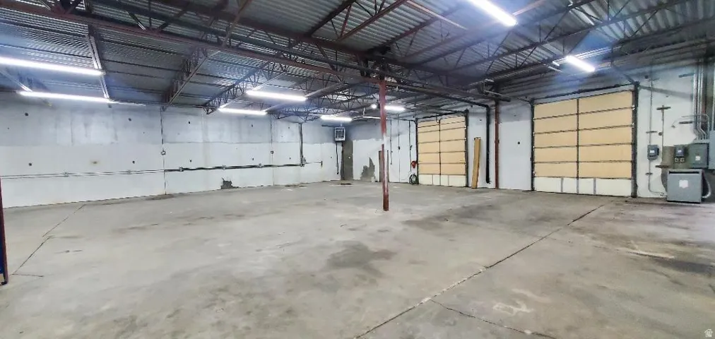 Spacious commercial interior featuring a concrete floor, exposed metal truss ceiling, fluorescent lighting, two large roll-up doors, and exposed block walls