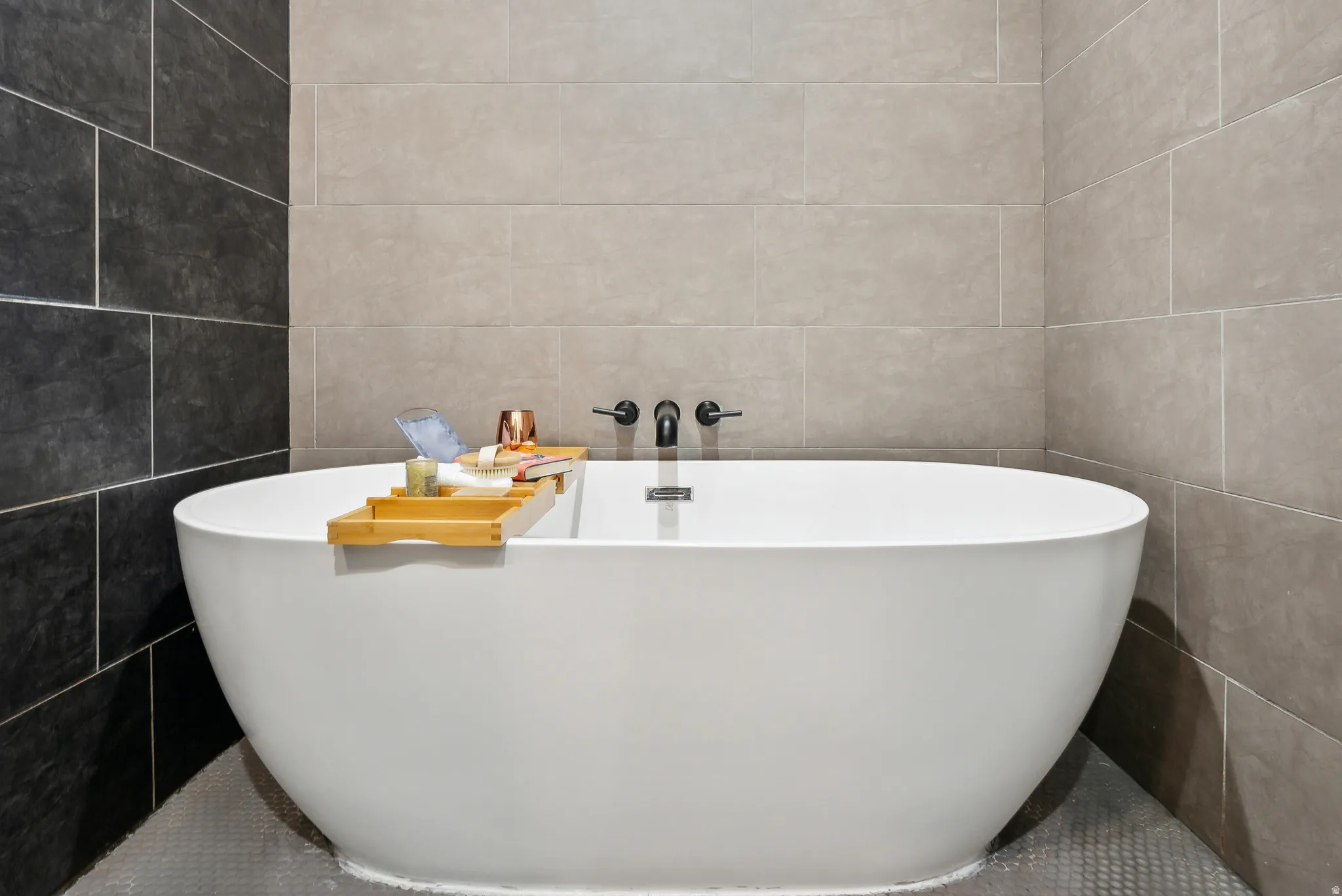 Freestanding soaking tub with matte black wall-mounted fixtures, complemented by large format gray and charcoal tile walls and penny tile flooring