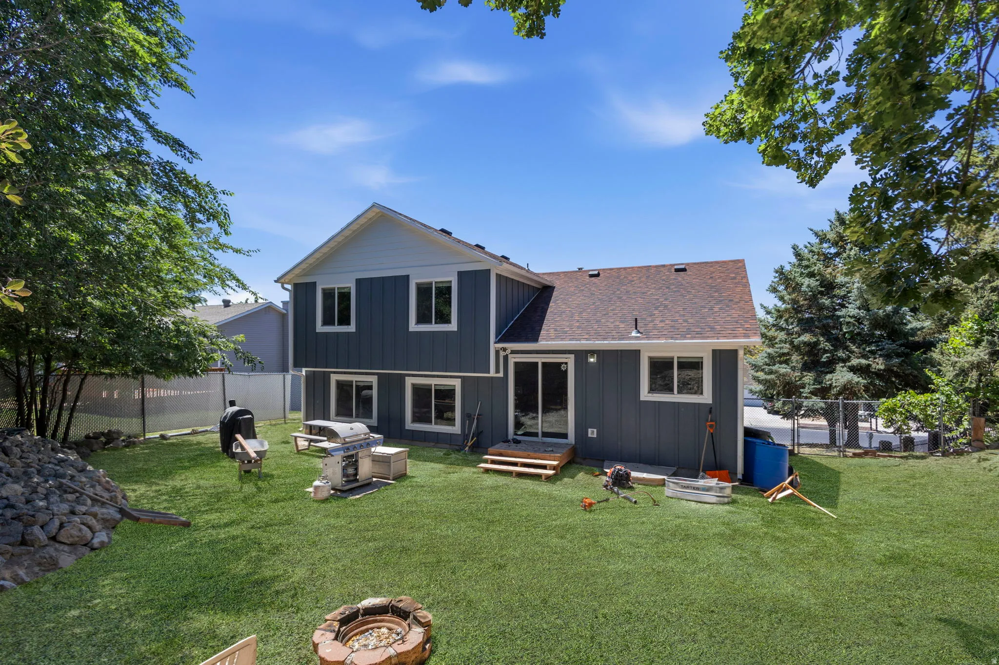 Expansive backyard featuring a manicured lawn, a built-in fire pit with stone surround, a multi-level structure with a sliding glass door, a rock garden, and mature trees