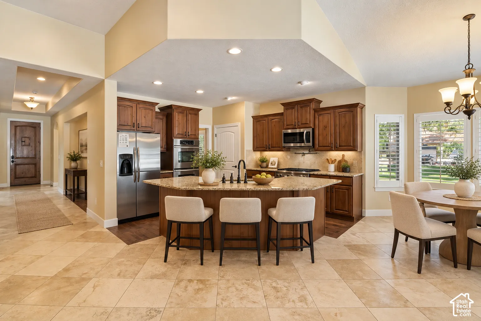 Kitchen featuring stained wood cabinetry, stone countertops, stainless steel appliances, a central island with an integrated sink, and stone tile flooring