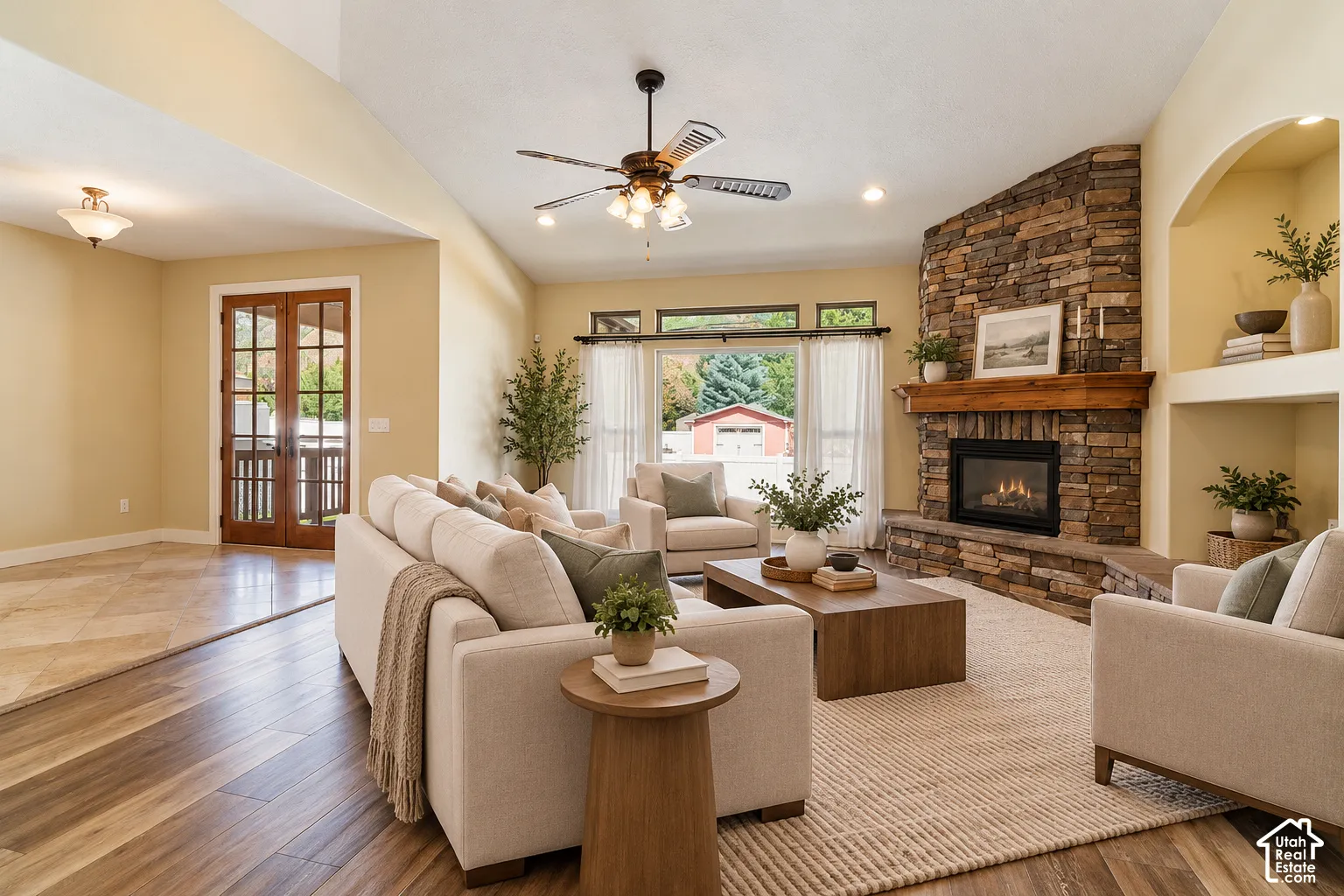 Vaulted ceiling living area featuring wood-finish flooring, a full-height stone fireplace with a prominent timber mantel, arched built-in shelving, and dual French doors