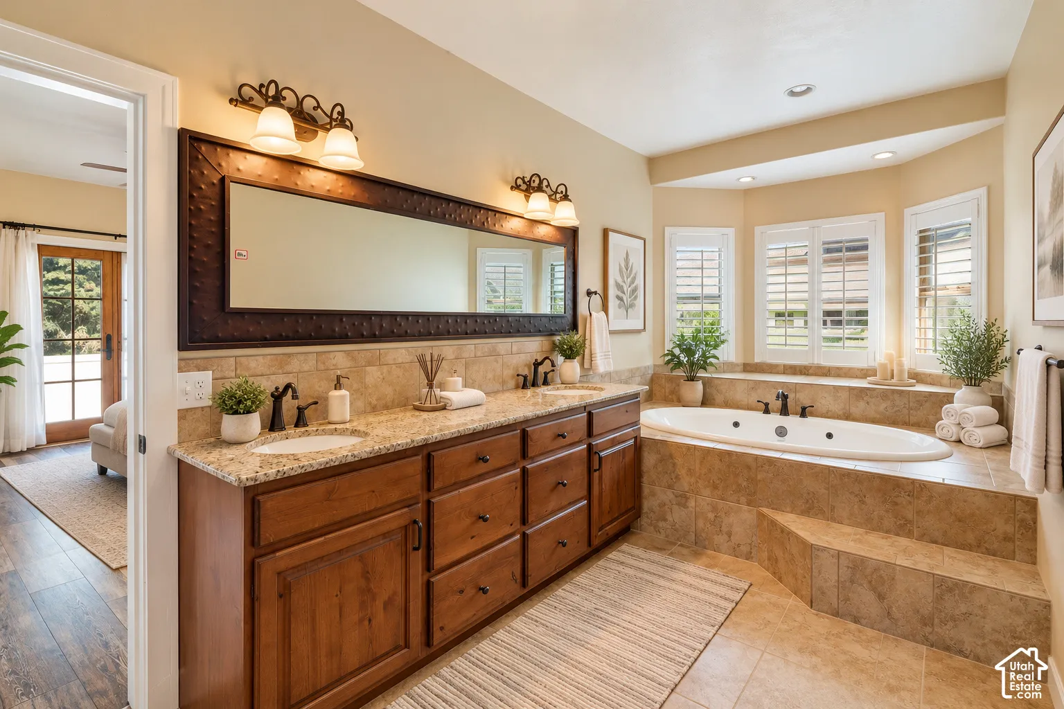 Dual-sink vanity with stone countertop and custom hammered-metal framed mirror, sunken jetted soaking tub, tiled tub surround, and plantation shutters