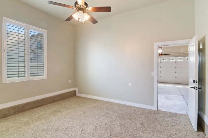 Carpeted interior space featuring a ceiling fan with light fixture, window with plantation shutters, light-toned walls, and white baseboards