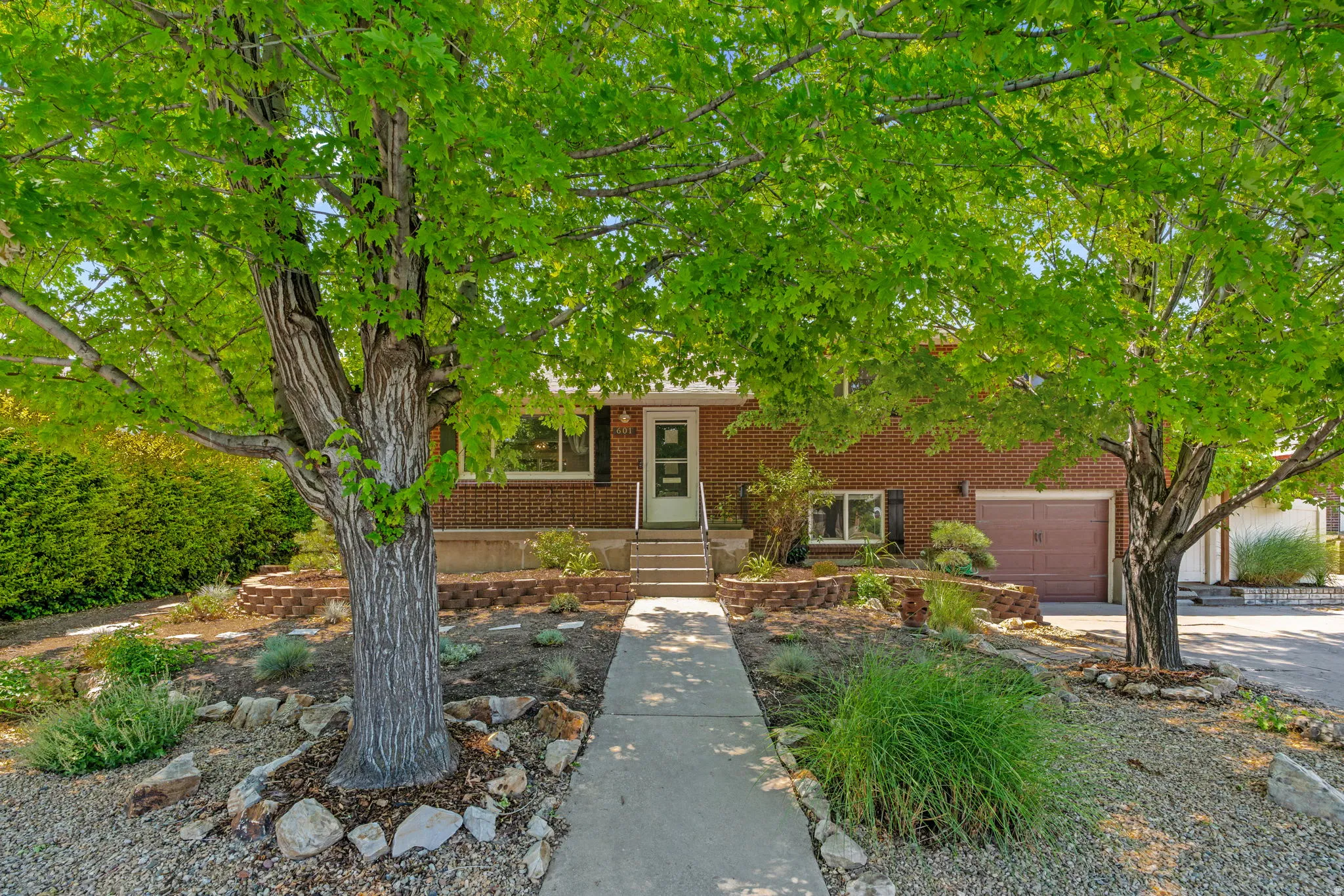 Brick facade residence featuring a covered entry, established landscaping with mature trees, a concrete pathway, and an attached one-car garage