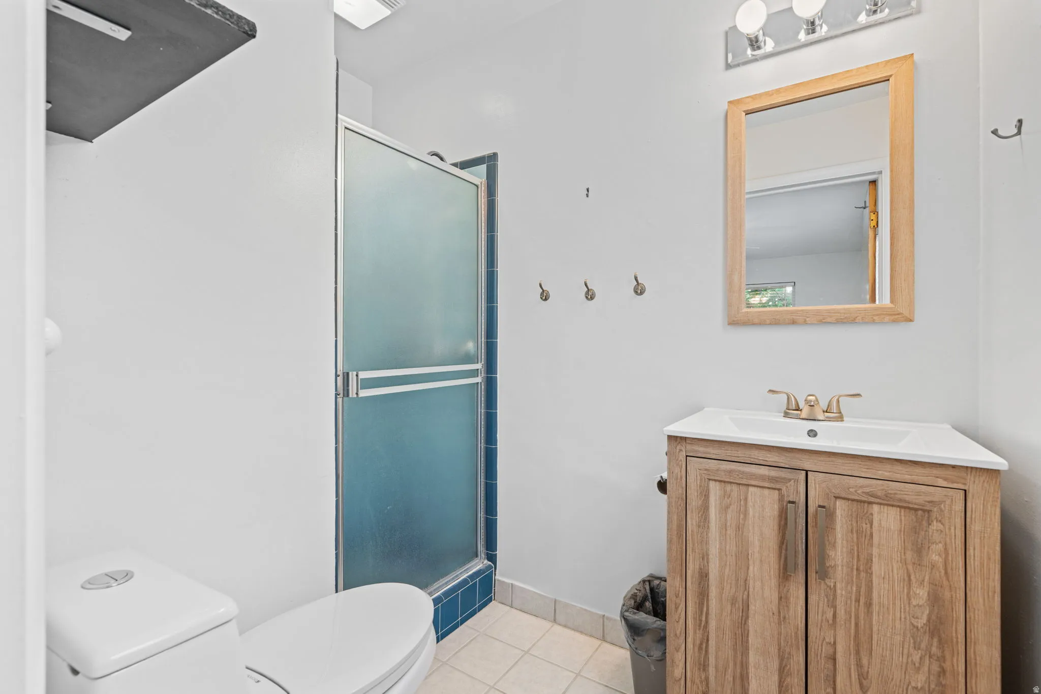 Bathroom featuring a wood-finish vanity with an integrated sink, a framed mirror, a three-bulb vanity light fixture, and a glass-enclosed shower with blue tile surround