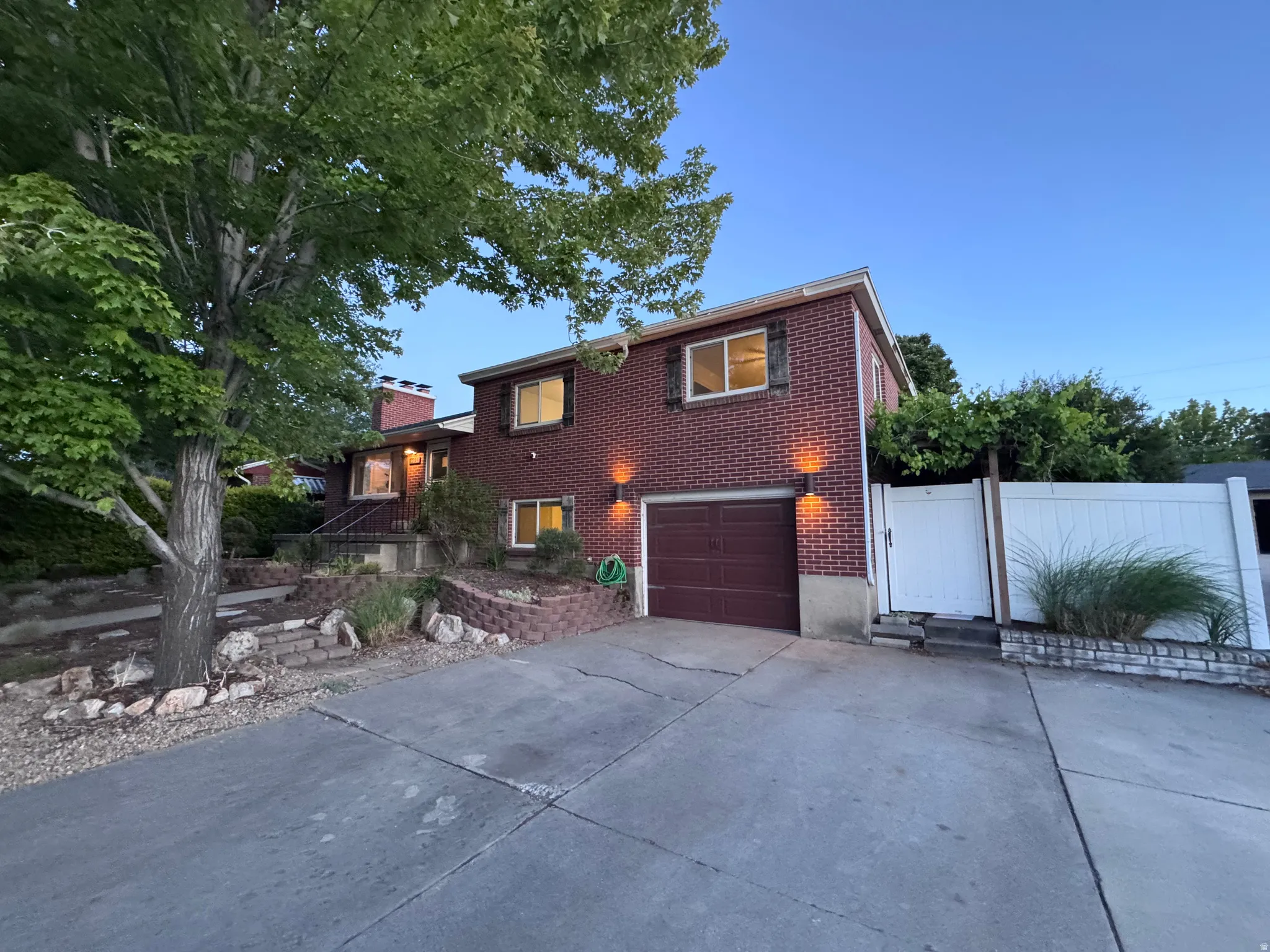 Driveway with RV parking, brick exterior featuring an attached garage, white privacy fence, multi-level landscaping with retaining walls, and mature trees