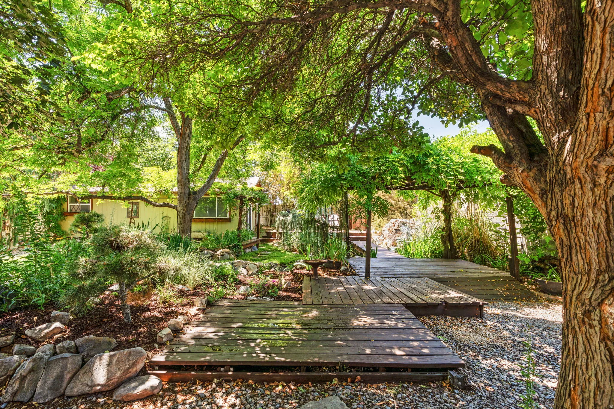 Expansive outdoor area featuring mature trees, a wooden deck, established landscaping, and natural stone elements