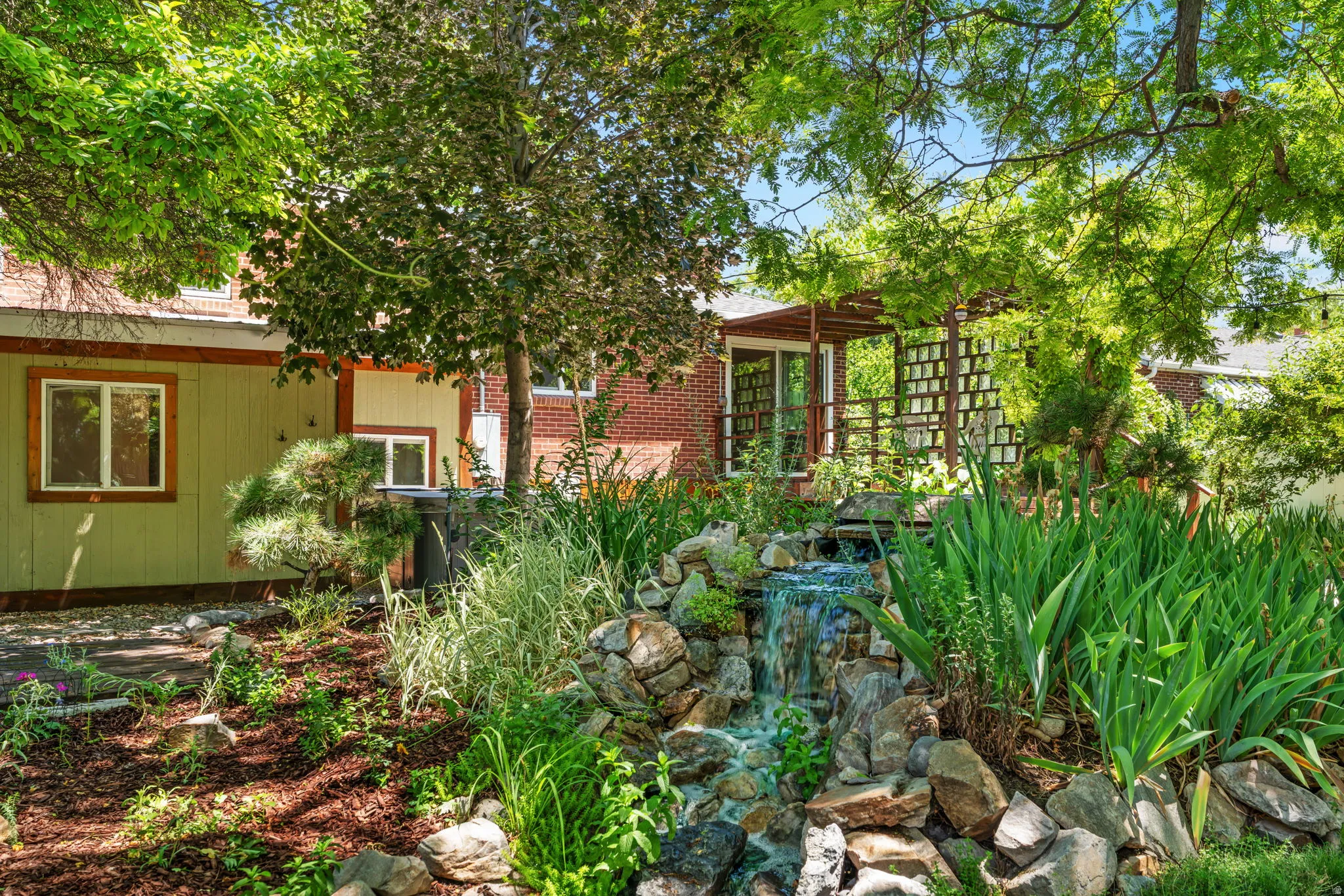 Lush landscaping featuring a multi-tiered rock waterfall, mature trees, and a wooden pergola with lattice detailing