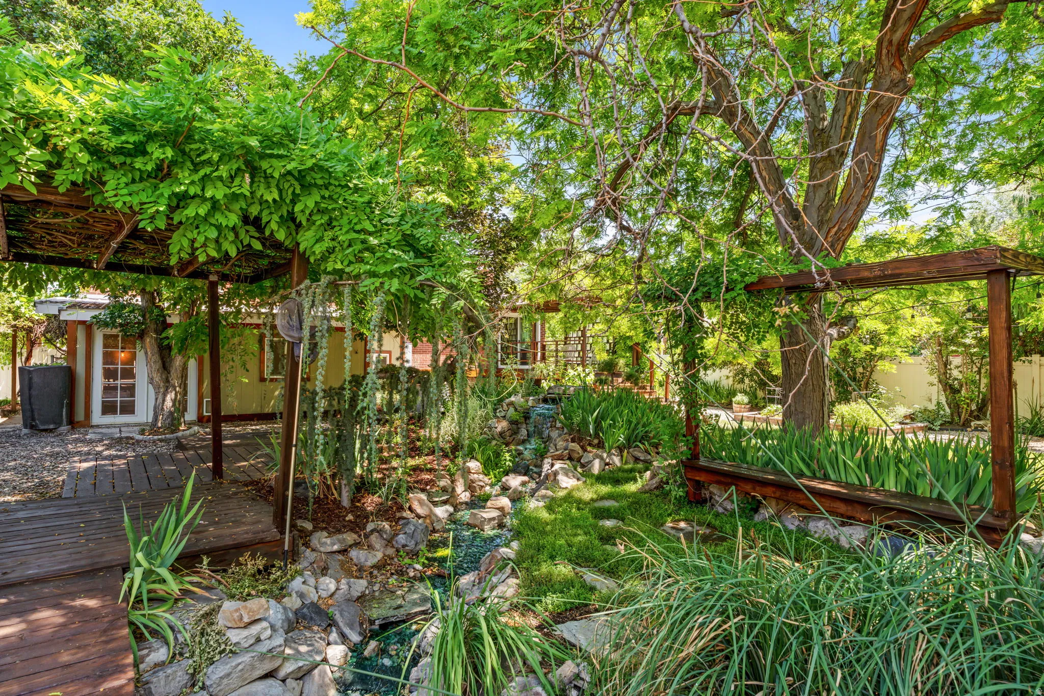 Lush backyard sanctuary featuring mature trees, thriving greenery, a wood-deck walkway, a rock-lined water feature, and a pergola with climbing vines