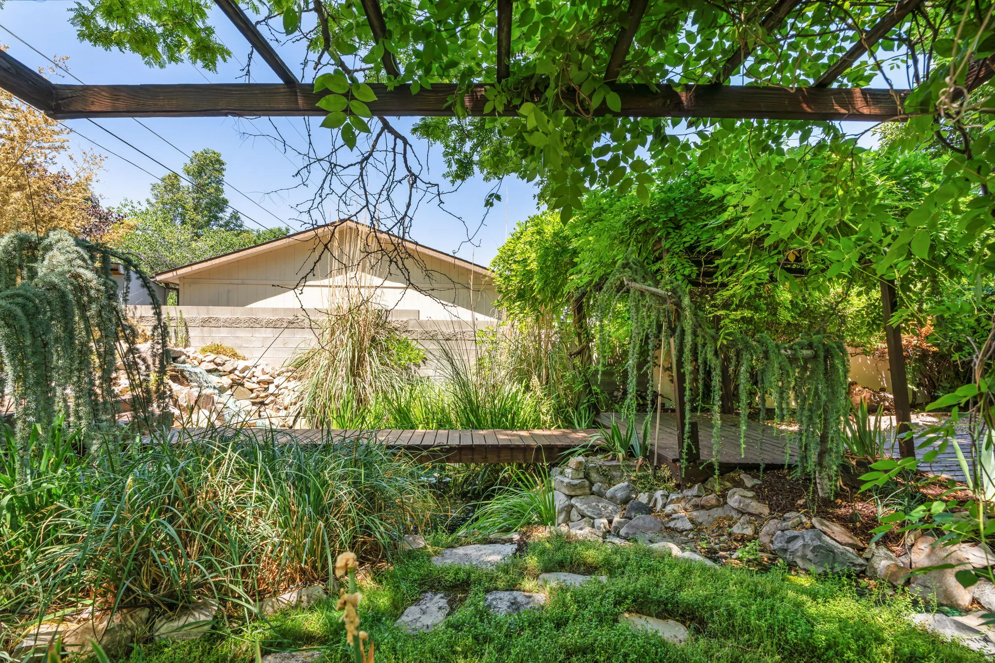 Lush garden landscaping featuring a wooden pergola, diverse plant varieties, a wooden footbridge, and natural rock elements