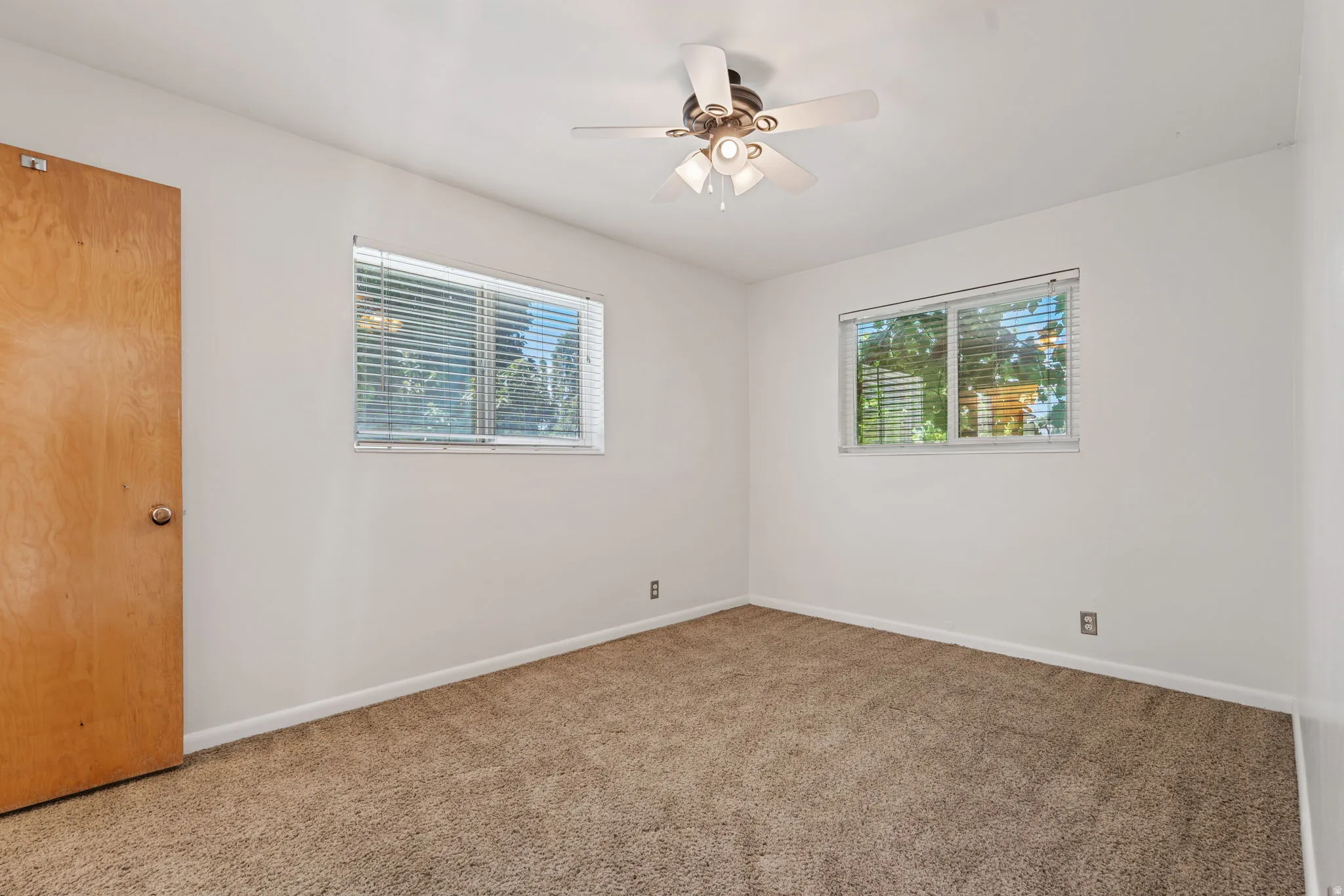 Neutral wall tones featuring textured carpeting, two windows with blinds, a ceiling fan with integrated lighting, and a wood-finish door