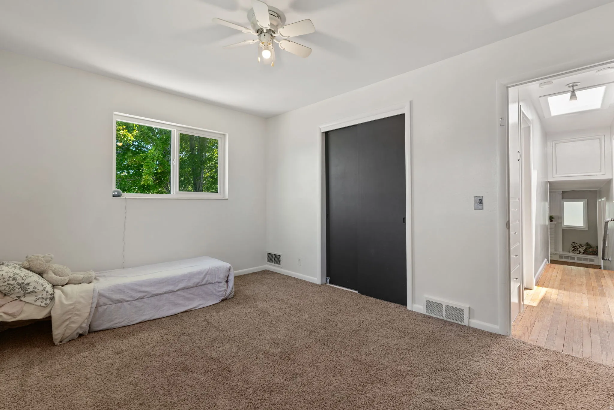 Carpeted room featuring a white ceiling fan with integrated lighting, a large window with green tree scenery, and a dark bi-fold closet