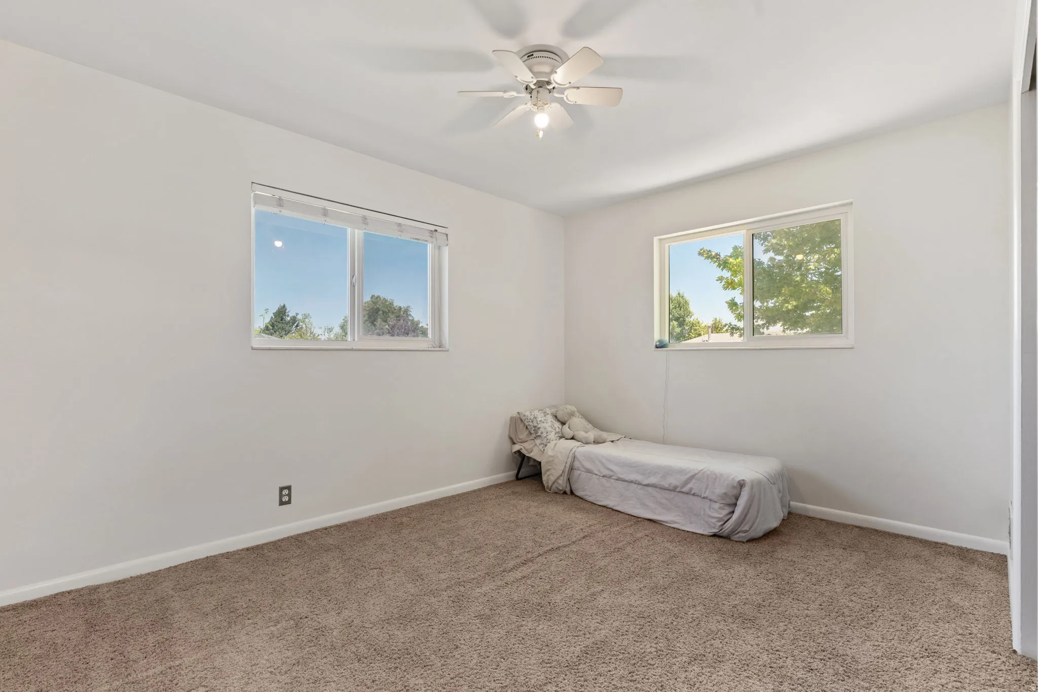 Carpeted room featuring white walls, baseboards, two windows with white frames, and a ceiling fan with integrated lighting