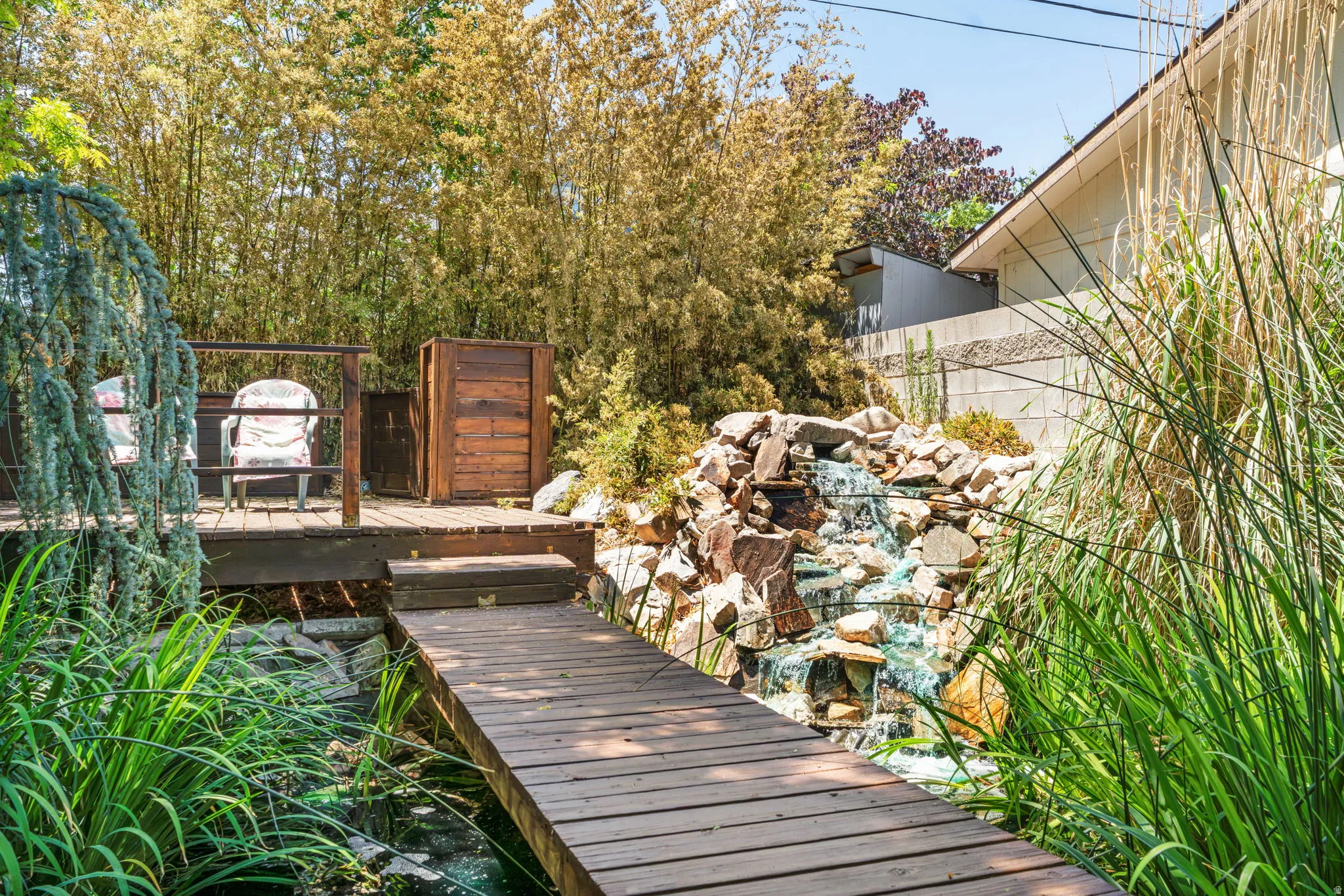 Expansive outdoor space featuring a wood-finish deck with railing, a decorative rock waterfall, a pond with a wood-finish bridge, and mature foliage