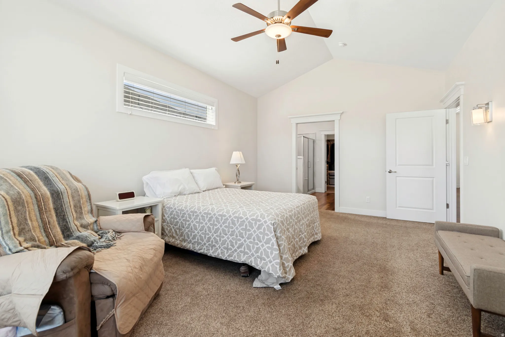 Spacious bedroom featuring vaulted ceilings, a ceiling fan with integrated lighting, soft carpeting, transom window with blinds, and white trim throughout