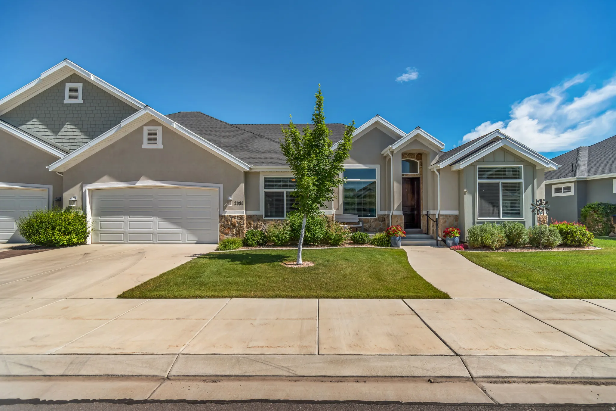 Stucco exterior with stone accents, featuring a two-car garage, mature landscaping, and a concrete walkway