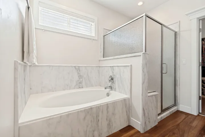 Bathroom featuring a built-in soaking tub with a polished chrome faucet, a glass-enclosed shower with a textured panel, and wood-finish flooring