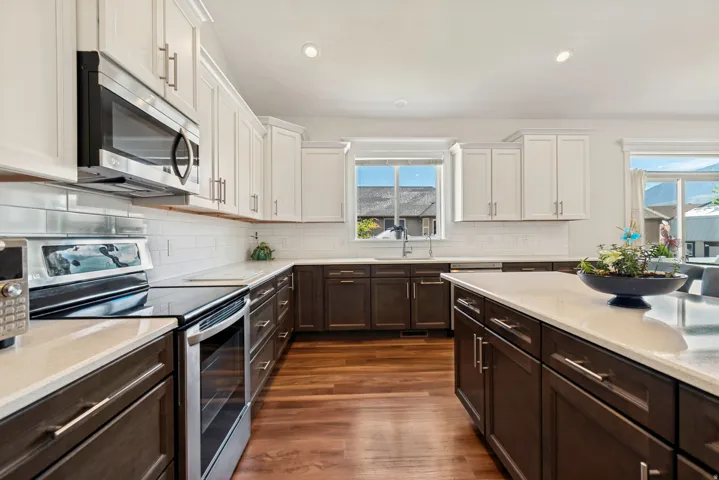 Modern kitchen featuring wood-finish flooring, two-tone cabinetry with white upper and dark lower cabinets, stainless steel appliances, and a central island with a light-toned countertop