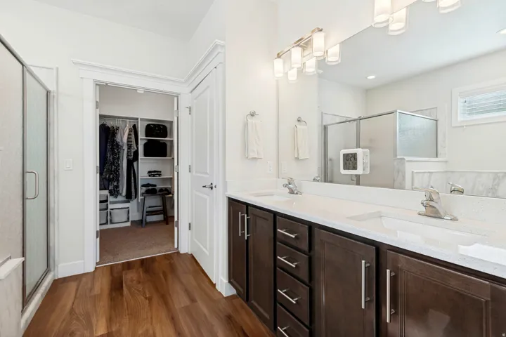 Dual vanity with integrated sinks, wood-finish flooring, a frameless glass shower enclosure, a large mirror, and a walk-in closet with built-in shelving