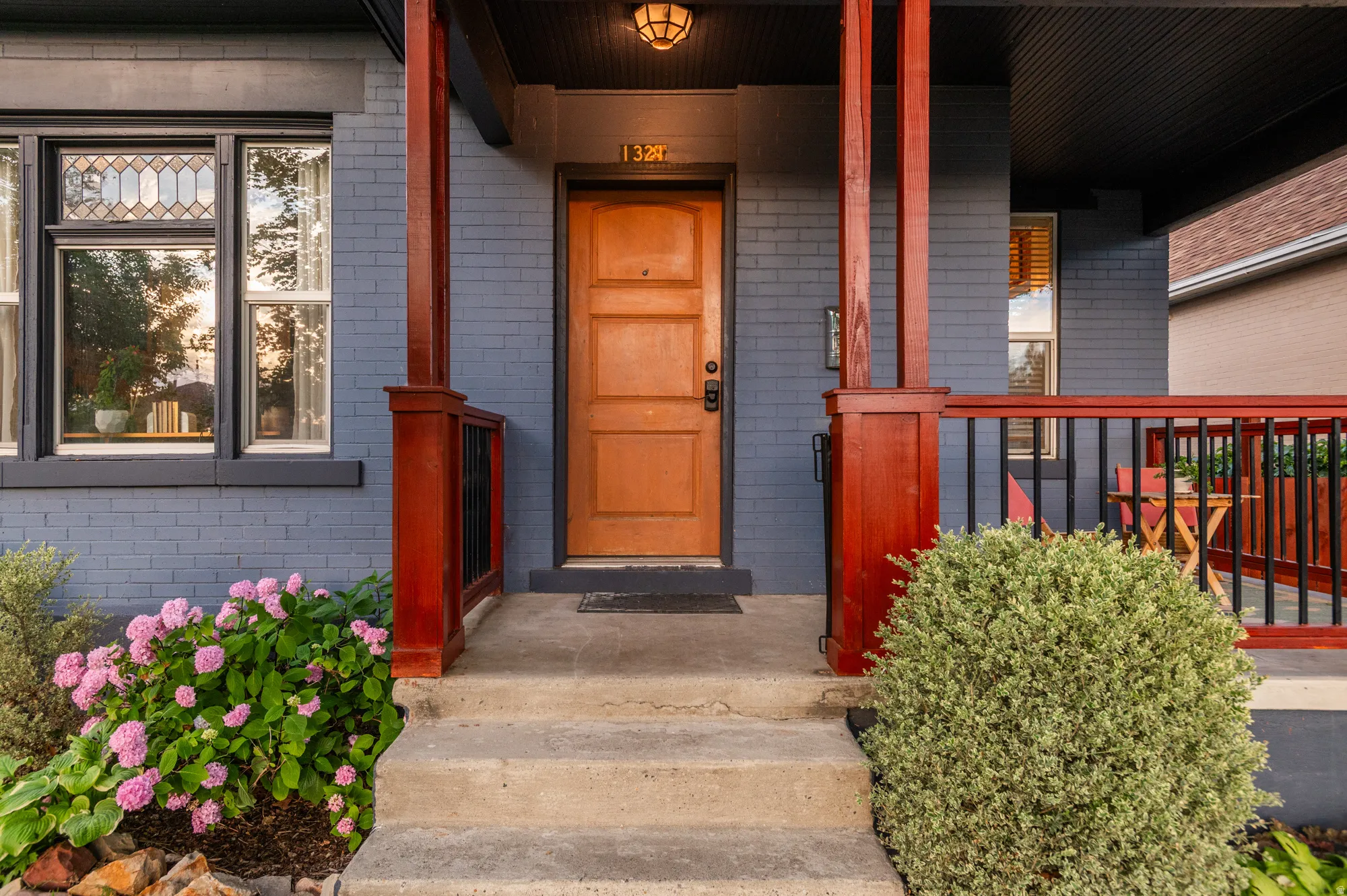 Brick exterior with a wooden front door, concrete steps, stained glass window accents, a covered porch with dark red pillars and railing, and established landscaping