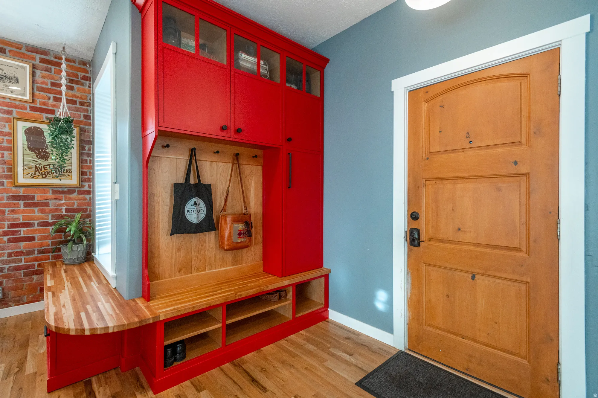 Entryway featuring a solid wood panel door, built-in red cabinetry with glass-front uppers, a wood-finish bench with integrated storage, and exposed brick accent wall