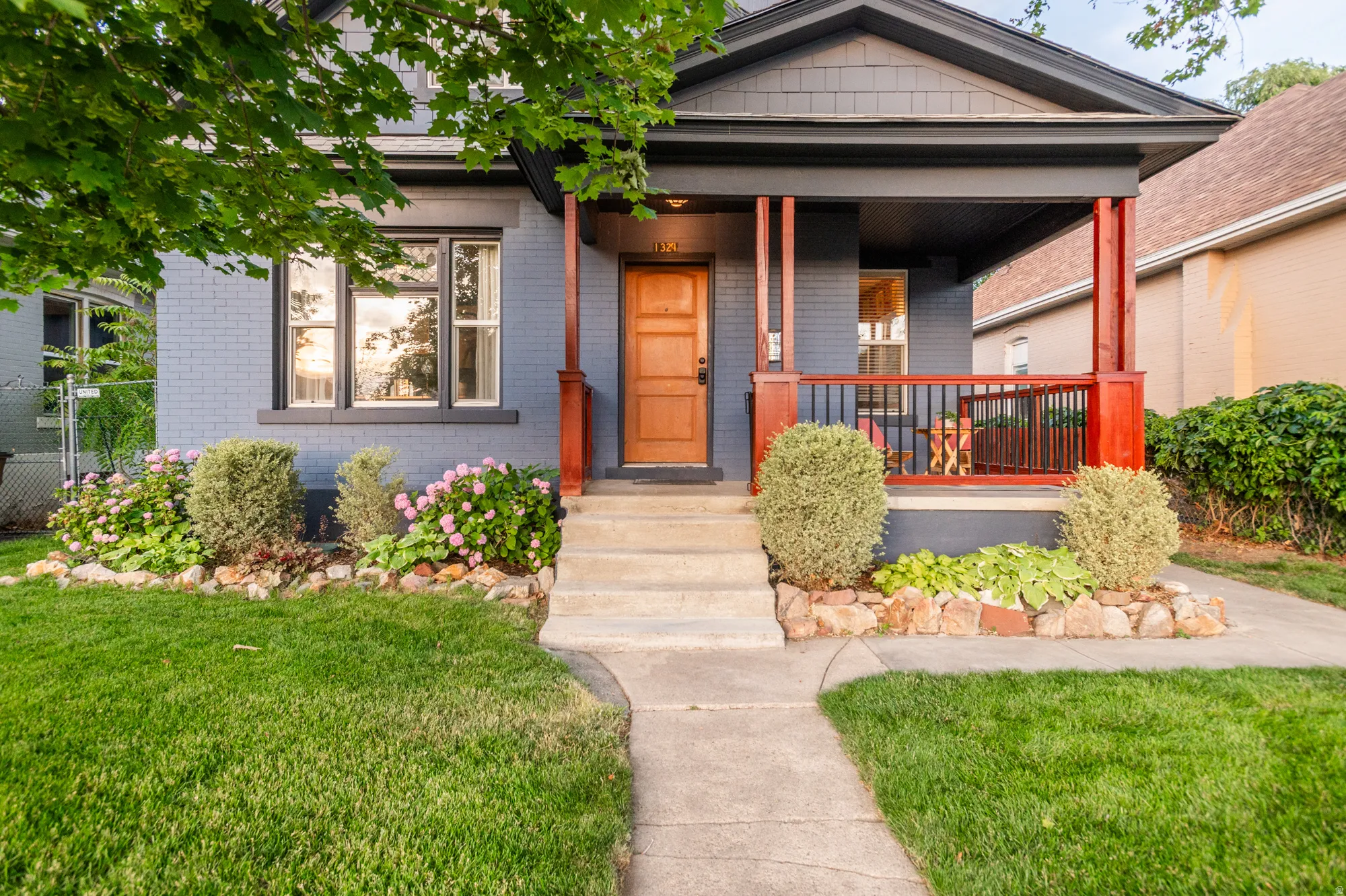 Brick exterior with painted finish and dark trim, featuring a covered porch with red-stained wood accents, a solid wood entry door, concrete steps, and landscaped front beds