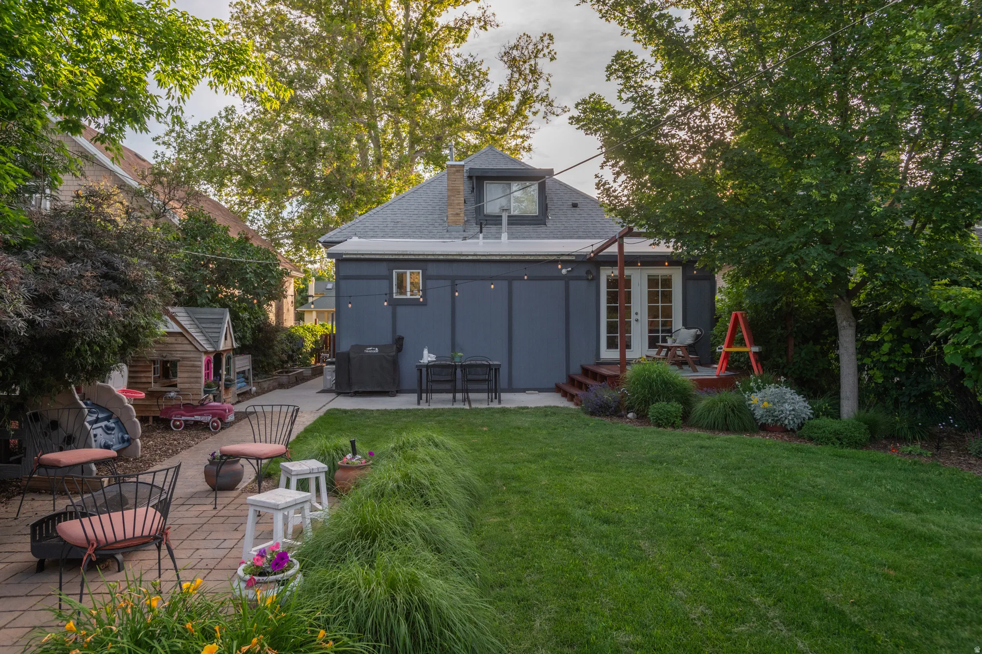 Rear exterior featuring a dormer window, French doors, a raised wooden deck, a brick paver patio, and mature landscaping with a manicured lawn