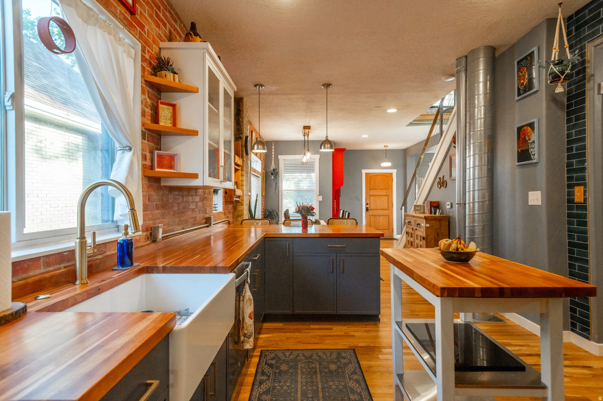Contemporary kitchen featuring wood-finish countertops, exposed brick backsplash, a farmhouse sink with a brass-finish faucet, dark base cabinetry, and overhead pendant lighting