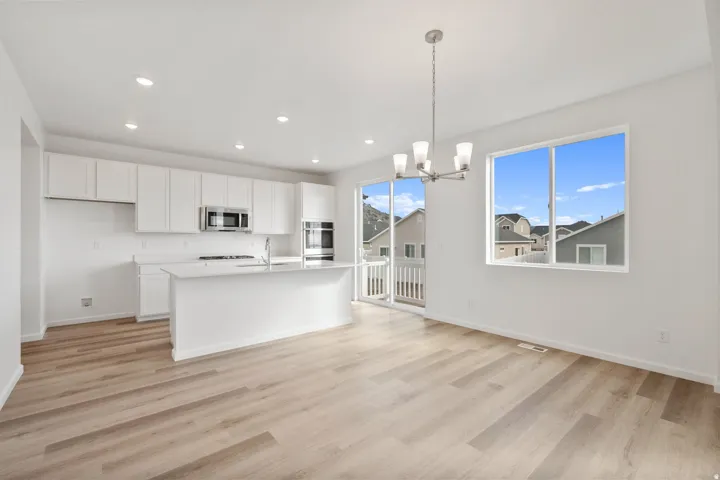 Open-concept kitchen and dining area featuring light wood-finish flooring, white cabinetry, a central island with a sink, recessed lighting, and stainless steel appliances including a microwave and double oven