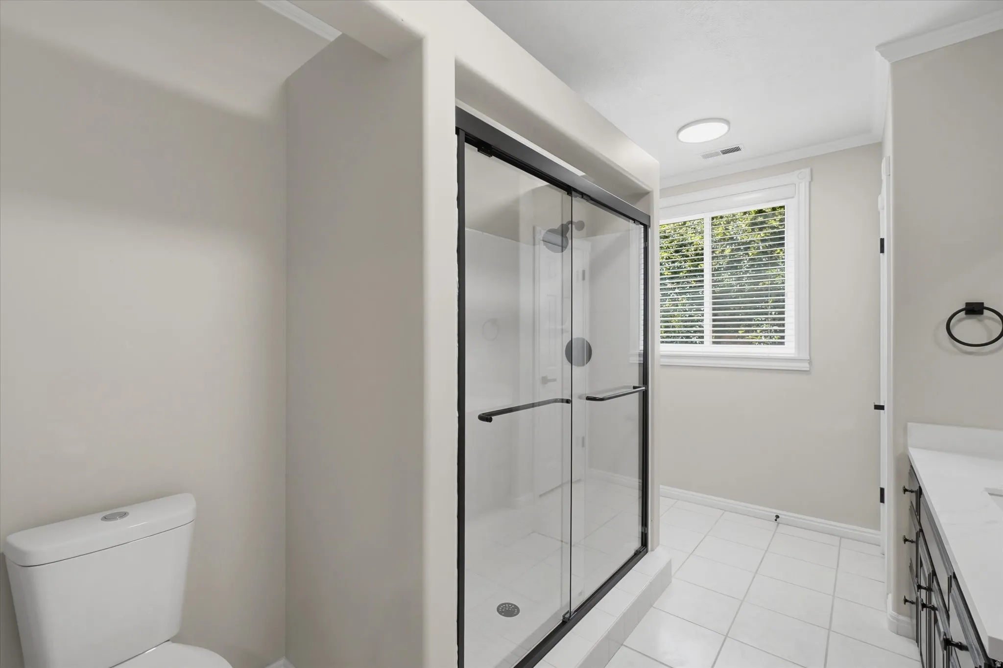 Full bathroom featuring a frameless glass shower enclosure with matte black trim, white square floor tiles, and a window with blinds