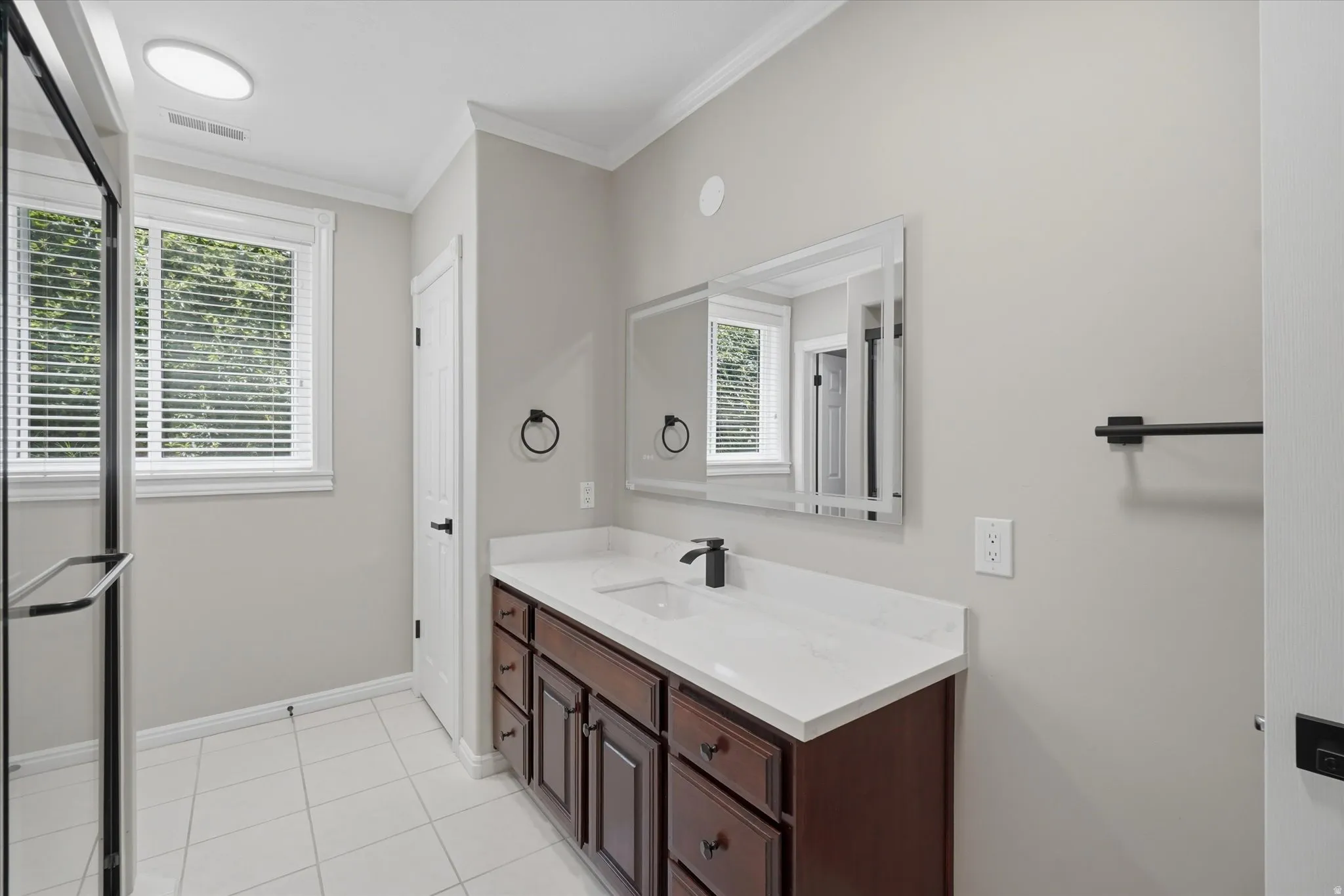 Bathroom featuring a rich wood-finish vanity with a white countertop and integrated sink, a frameless glass shower enclosure, a rectangular wall-mounted mirror, and tile flooring