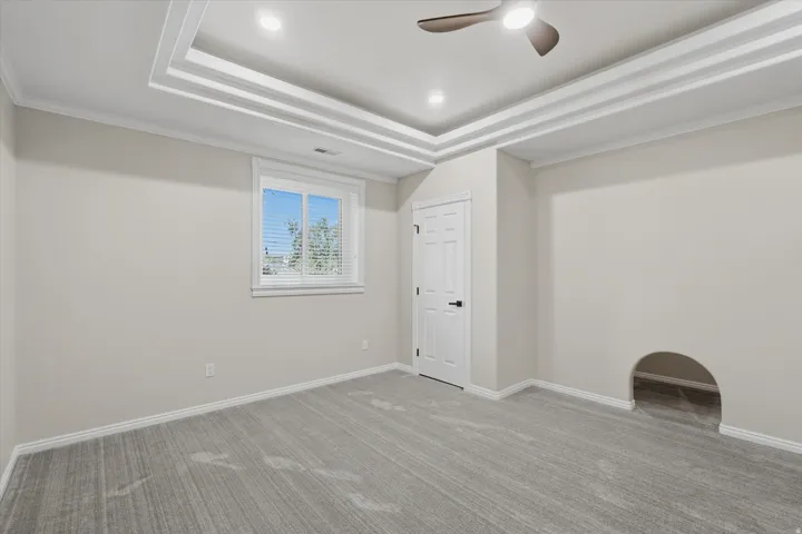 Recessed tray ceiling with integrated lighting, modern ceiling fan, gray carpet flooring, white trim, and a window with blinds
